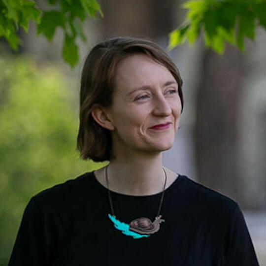 A headshot of Courtney with short light brown almost auburn hair, a black shirt, and a snail necklace.