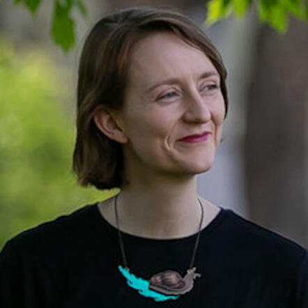 A photo of Courtney, a white person with short auburn hair wearing a snail necklace, looking away from the camera and smiling.
